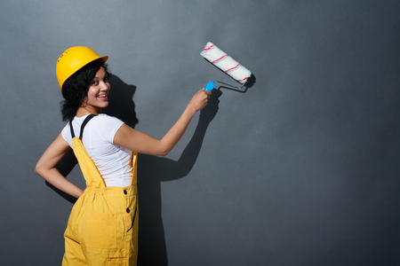 Happy smiling mixed race construction woman wearing yellow protect helmet and overall adjusting helmet with a painting roller going to paint a grey wallの写真素材