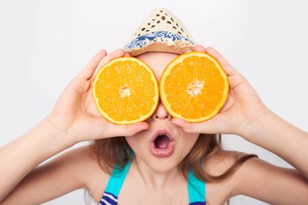 Llittle girl in swimsuit and summer hat having fun with half oranges making fake eyeglasses with expression of surprise, search conceptの写真素材