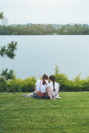 Two girls friends sitting on grass in a park chatting, back viewの写真素材
