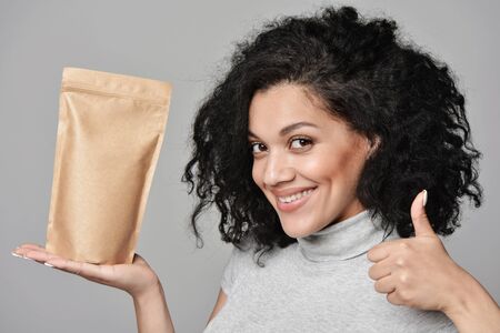 Smiling woman showing craft paper pouch bag with copy space and gesturing thumb up, closeup portrait over grey background.の写真素材