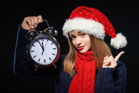 Closeup portrait of smiling girl wearing Santa hat holding big alarm clock time approaching midnight and pointing to the side at copy space, over dark backgroundの写真素材