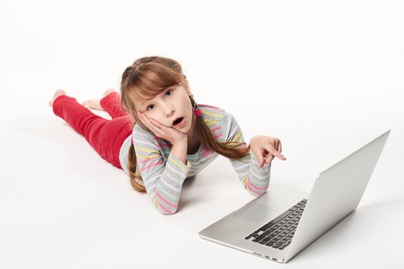 Surprised little girl lying on stomach on the floor with laptop, pointing at screen with amazement, over white backgroundの写真素材