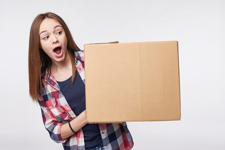 Delivery, relocation and unpacking. Excited young woman holding cardboard box, looking at blank copy space at box, surprised, isolatedの写真素材