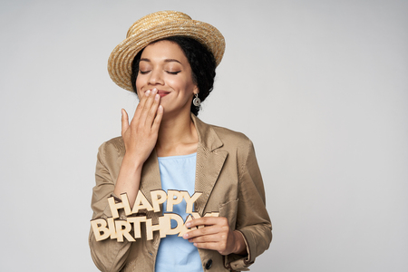 Beautiful smiling mixed race woman holding happy birthday text excited of happiness with closed eyes covering her mouth with hand, isolated on gray backgroundの写真素材
