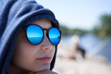 Closeup of pensive little girl in a hood and sunglasses looking away in thoughts with reflection of river in glassesの写真素材
