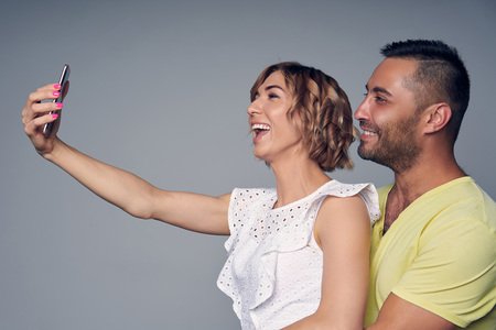 Happy couple in studio laughing making self portrait on smart phone, over grey backgroundの写真素材