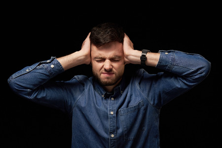 Man with problems. Young man in denim shirt holding head in hands, frowning, isolated on black...