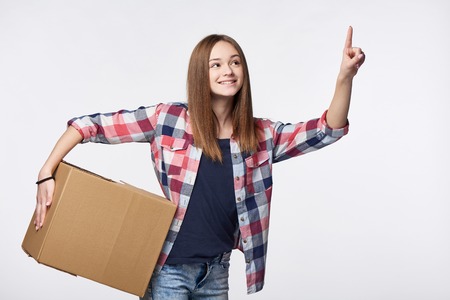 Delivery, relocation and unpacking. Smiling young woman holding cardboard box and pointing finger up or pushing imaginary buttonの写真素材