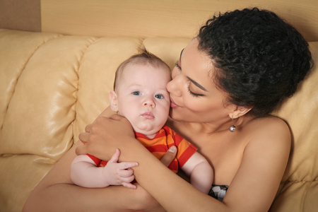 Pretty mixed race african american - caucasian woman holding her caucasian baby in arms kissing sitting on sofaの写真素材