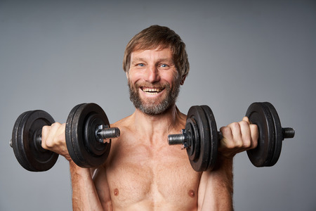 Portrait of healthy mature man shirtless standing with dumbbells looking at camera, over grey studio backgroundの写真素材