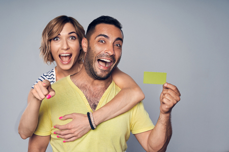 Happy surprised couple embracing, looking at camera showing blank credit card and pointing at you, over grey backgroundの写真素材