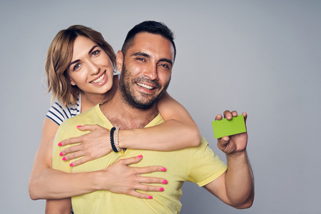 Happy couple embracing, looking at camera showing blank credit card, over grey backgroundの写真素材