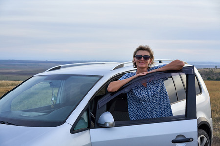 Woman standing leaning on a car door looking at camera smiling, on countryside on twilightの写真素材