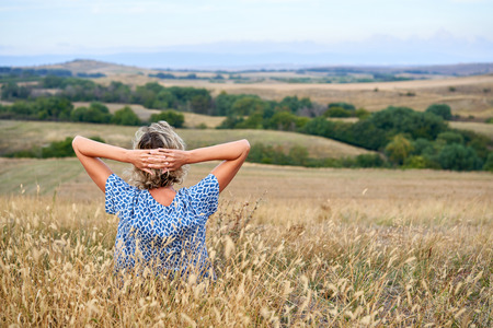 Back view of a woman sitting in dry barren grass, looking in the horizon, relaxing with hands over headの写真素材
