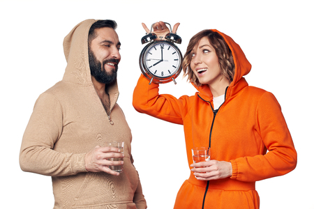 Young couple in home clothes standing on white background holding glasses of clear water showing big alarm clockの写真素材