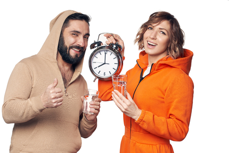 Young couple in home clothes standing on white background holding glasses of clear water showing big alarm clockの写真素材