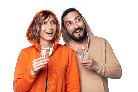 Young couple in home clothes standing on white background holding glasses of clear water looking to side at blank copy spaceの写真素材