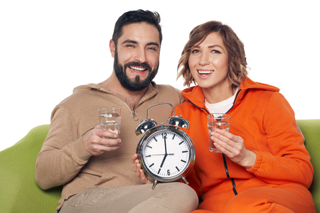 Young couple in home clothes sitting on sofa, holding glasses of clear water showing big alarm clockの写真素材