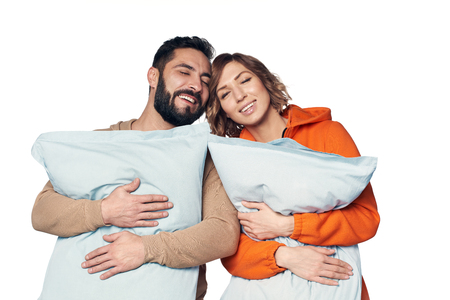 Happy young couple in home clothes hugging pillows enjoying with closed eyes, over white backgroundの写真素材