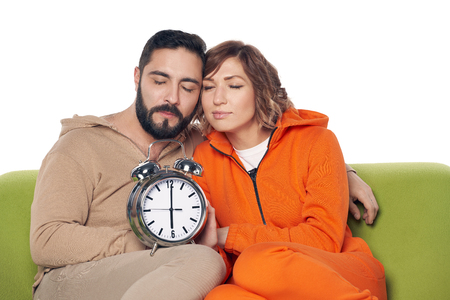Tired sleepy young couple in home clothes sitting on sofa holding big alarm clockの写真素材
