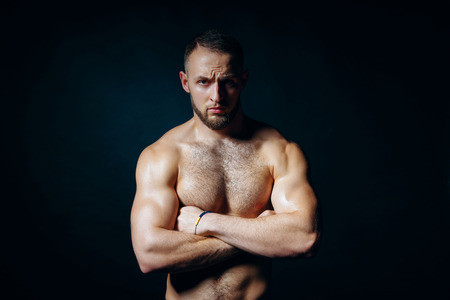 Low key portrait of muscular shirtless man at dark background standing with folded hands, looking at camera seriouslyの写真素材