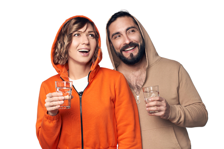 Happy smiling young couple in home clothes standing on white background holding glasses of clear water looking to side at blank copy spaceの写真素材