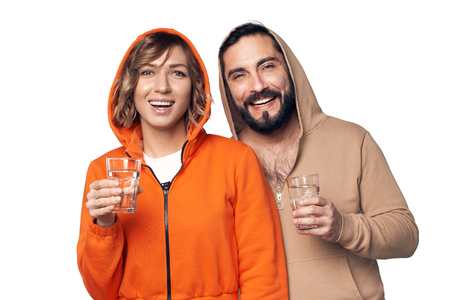 Happy young couple in home clothes standing on white background holding glasses of clear waterの写真素材
