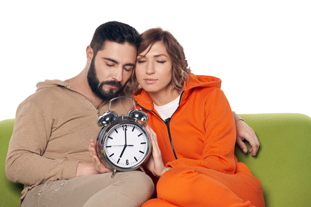 Tired sleepy young couple in home clothes sitting on sofa with closed eyes, with big alarm clock on kneesの写真素材