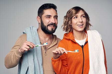 Morning hygiene. Young couple in lounge clothes with home towels over shoulder holding toothbrushes with toothpaste, looking to side smilingの写真素材
