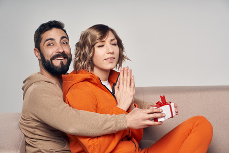 Holiday, celebration and family . Smiling man surprising his girlfriend with present at home sitting on sofaの写真素材