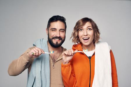 Morning hygiene. Young couple in lounge clothes with home towels over shoulder holding toothbrushes with toothpaste, smiling at cameraの写真素材
