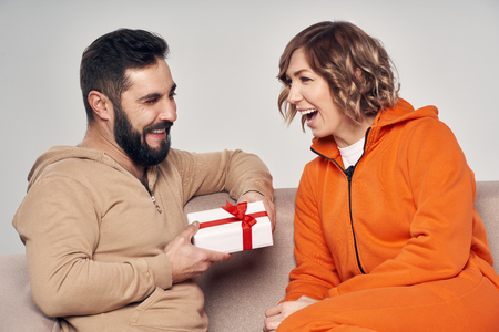 Holiday, celebration and family . Smiling man surprising his girlfriend with present at home sitting on sofaの写真素材