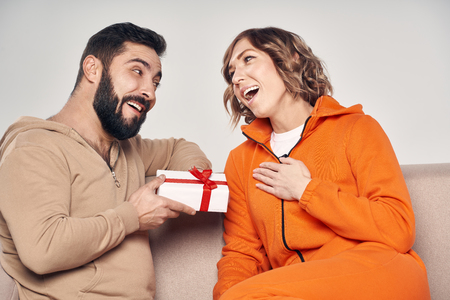 Holiday, celebration and family . Smiling man surprising his girlfriend with present at home sitting on sofaの写真素材