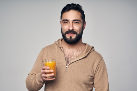 Young handsome bearded man holding a glass of orange juice isolated on grey backgroundの写真素材