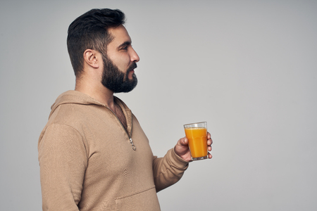 Profile of young handsome bearded man holding a glass of orange juice isolated on grey backgroundの写真素材