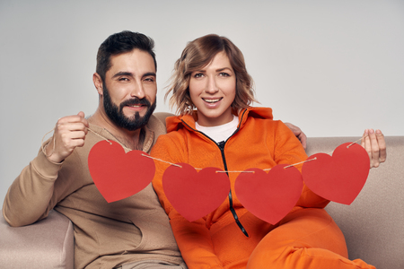 Love and family. Smiling couple sitting on sofa holding four heart shape garlandの写真素材