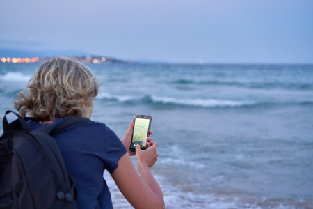 Woman taking a photo of a sea on smart phone on sunset at the beach, back view. Focus on phoneの写真素材