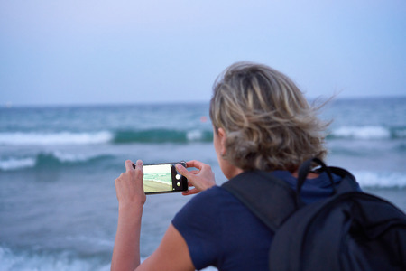 Woman taking a photo of a sea on smart phone on sunset at the beach, back view. Focus on phoneの写真素材