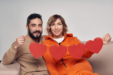 Love and family. Smiling couple sitting on sofa holding four heart shape garlandの写真素材