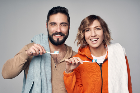 Morning hygiene. Young couple in lounge clothes with home towels over shoulder holding toothbrushes with toothpaste, smiling at cameraの写真素材