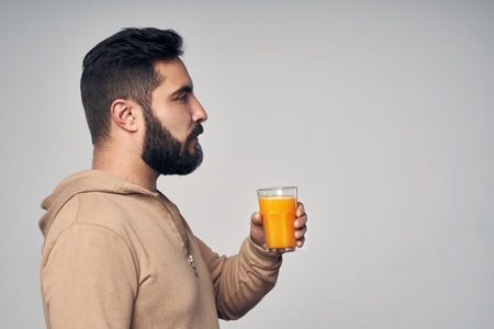 Profile of young handsome bearded man holding a glass of orange juice isolated on grey backgroundの写真素材