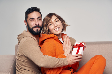 Holiday, celebration and family . Smiling man surprising his girlfriend with present at home sitting on sofaの写真素材