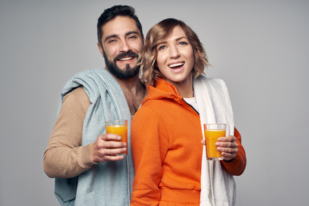 Young couple with towels over shoulder holding glasses of orange juice, isolated on grey backgroundの写真素材