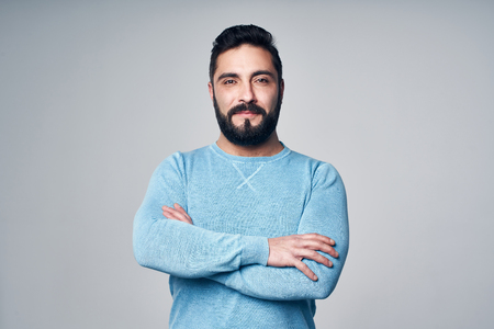 Confident Hispanic man in blue pullover standing with folded hands, looking at camera smilingの写真素材