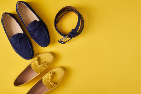 Dark blue suede man's and yellow woman's moccasins shoes with a belt over yellow background, top viewの写真素材