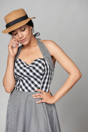 Pensive mixed race african american - caucasian woman in chequered summer dress and canotier straw hat standing with hand on cheek, looking down in thoughts, studio portraitの写真素材