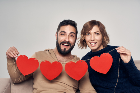 Love and family. Excited couple sitting on sofa holding four heart shape garland looking at cameraの写真素材