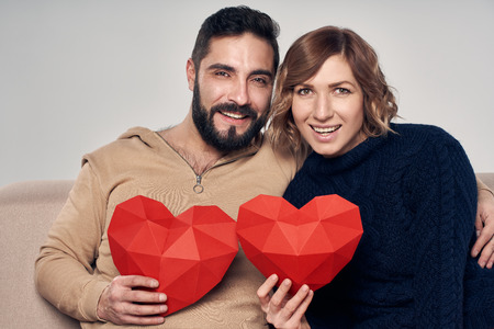 Love and family. Happy couple sitting on sofa holding heart shape looking at cameraの写真素材