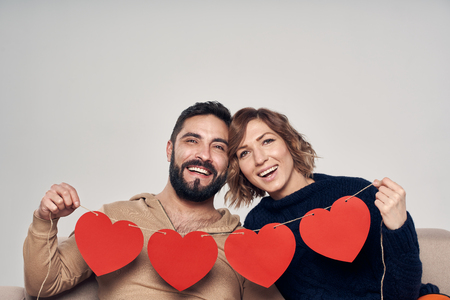 Love and family. Happy smiling couple sitting on sofa holding four heart shape garland looking at cameraの写真素材