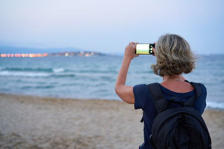 Woman taking a photo of a sea on smart phone on sunset at the beach, back view. Focus on womanの写真素材